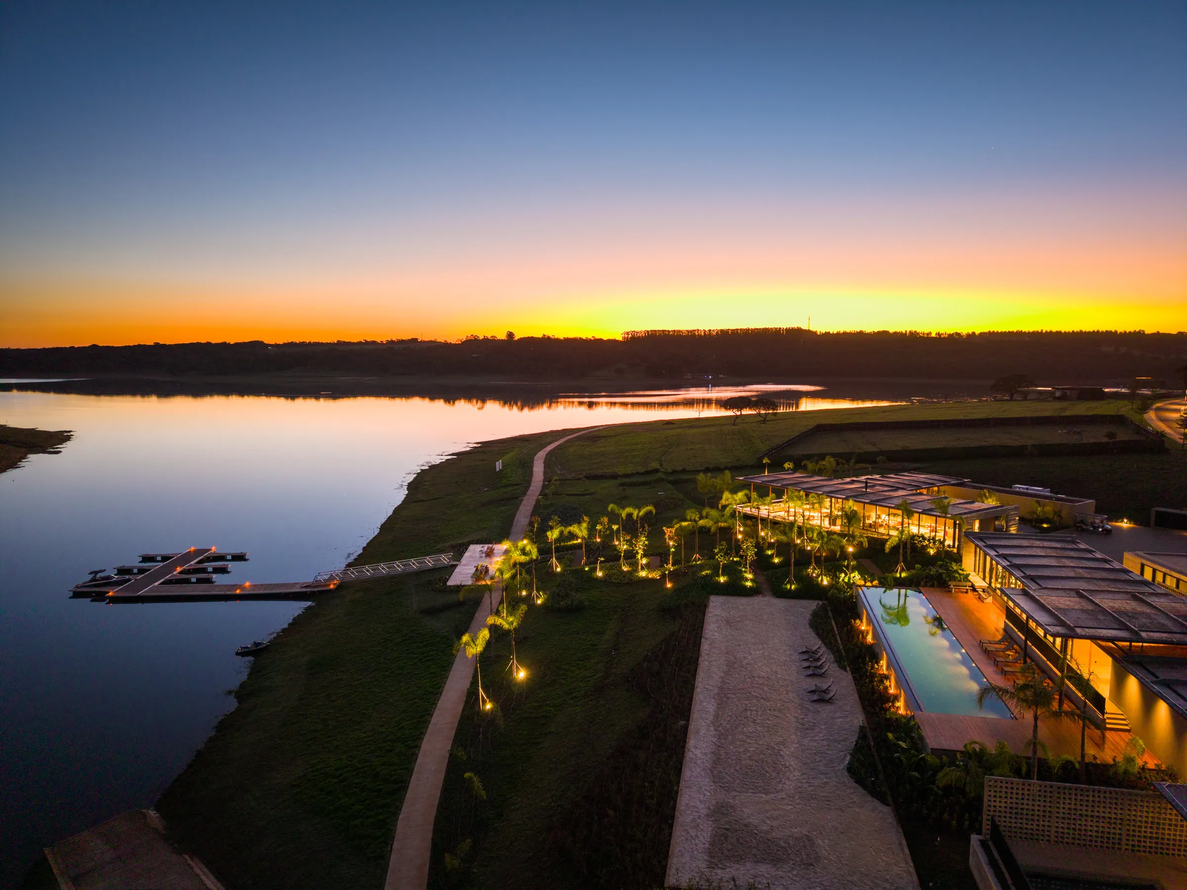 O clube iluminado ao anoitecer, com a piscina, o píer e a represa refletindo o pôr do sol